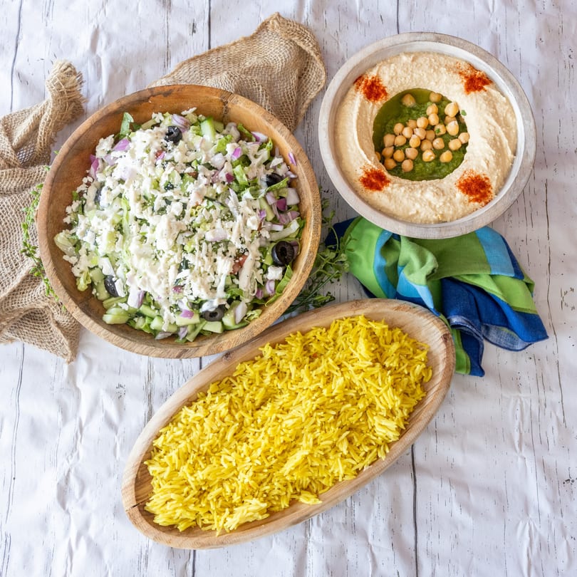 Overhead view of a salad, hummus with chickpeas, and yellow rice on a white crinkled surface.