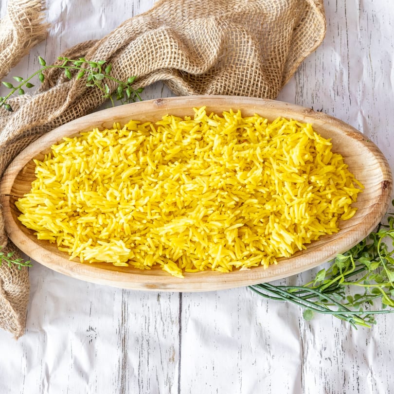 Bright yellow rice in a wooden bowl, with burlap and green sprigs on a white wooden surface.