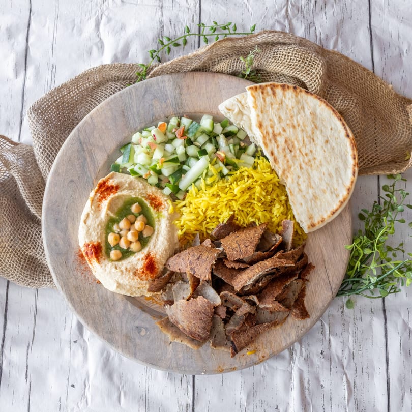 A Mediterranean platter with gyro meat, yellow rice, hummus, cucumber salad, and pita bread.