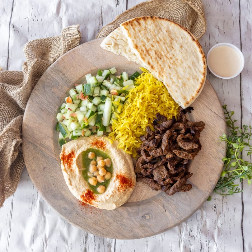 A wooden plate with beef shawarma, yellow rice, cucumber salad, hummus, pita bread, and sauce.