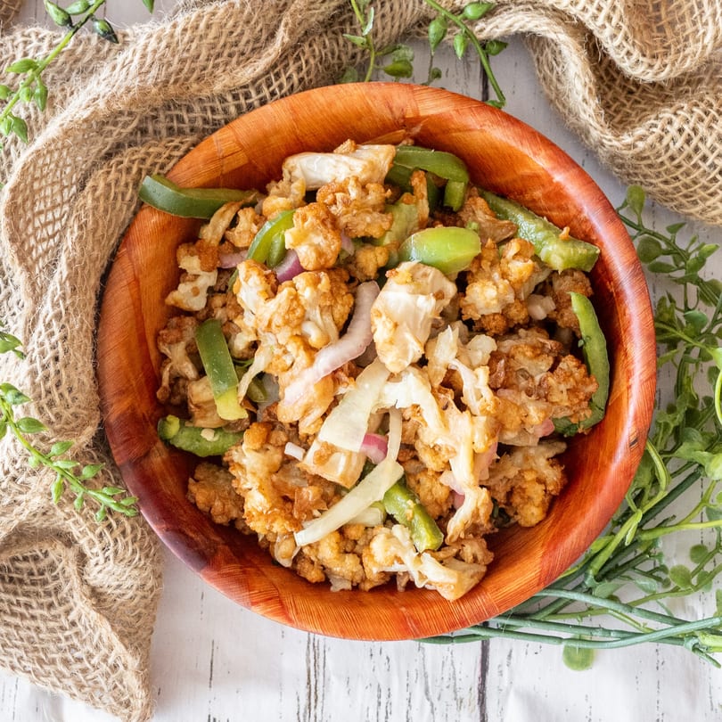 A wooden bowl filled with fried cauliflower, green peppers, and onions on a burlap cloth.