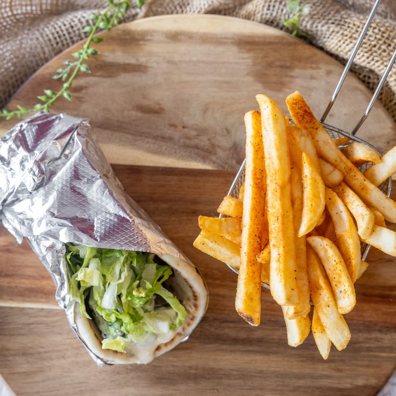 A foil-wrapped gyro with lettuce and a metal basket of seasoned french fries on a wooden board.