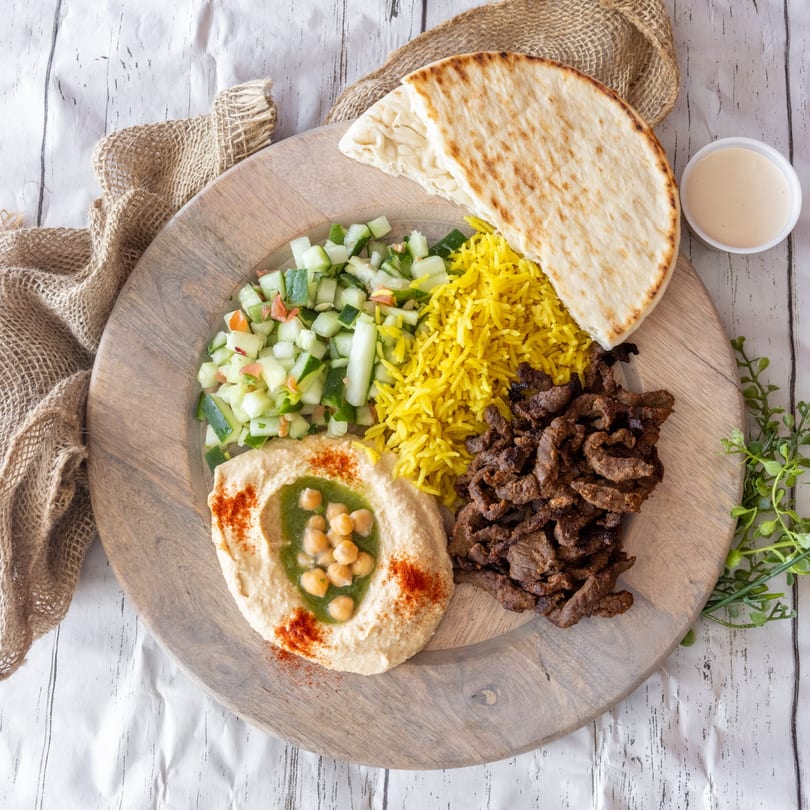 Shawarma plate with meat, yellow rice, cucumber salad, hummus, pita bread, and a side of sauce.