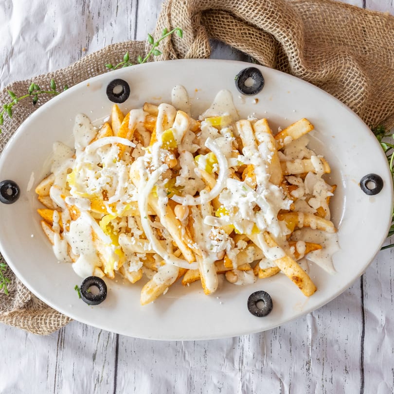 A white oval plate of loaded fries with white sauce, cheese, onions, and peppers, garnished with olives.