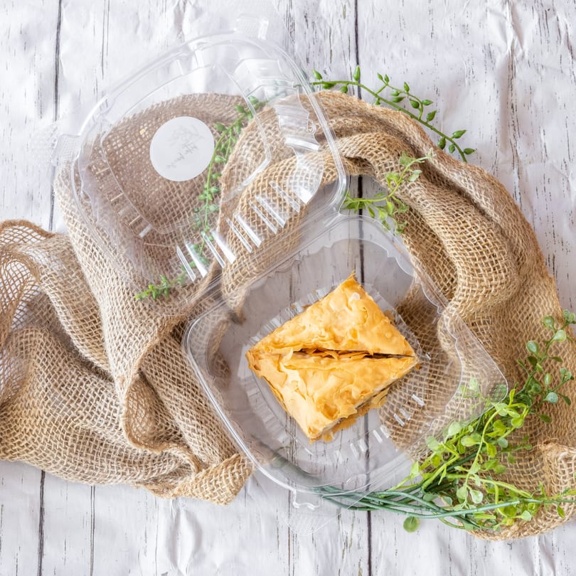 A triangular pastry in a clear plastic container, on burlap with green sprigs, on a white wooden surface.