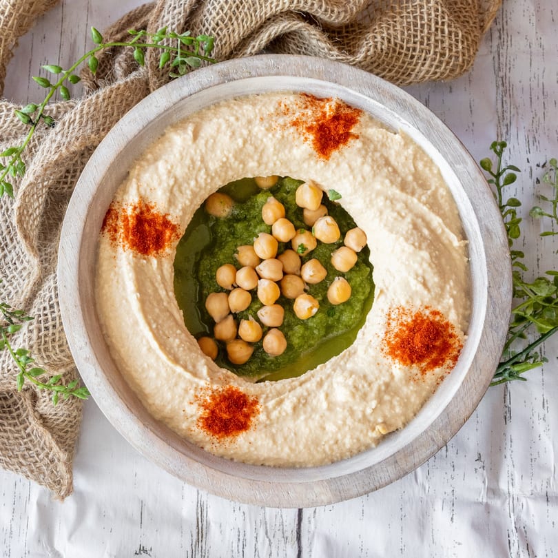 Creamy hummus with a green pesto center, topped with chickpeas and paprika, on a rustic wooden plate.