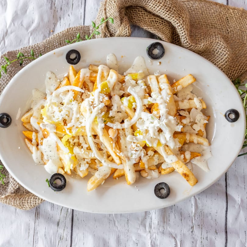 A white oval platter of golden fries topped with creamy sauce, crumbled cheese, onions, and black olives.