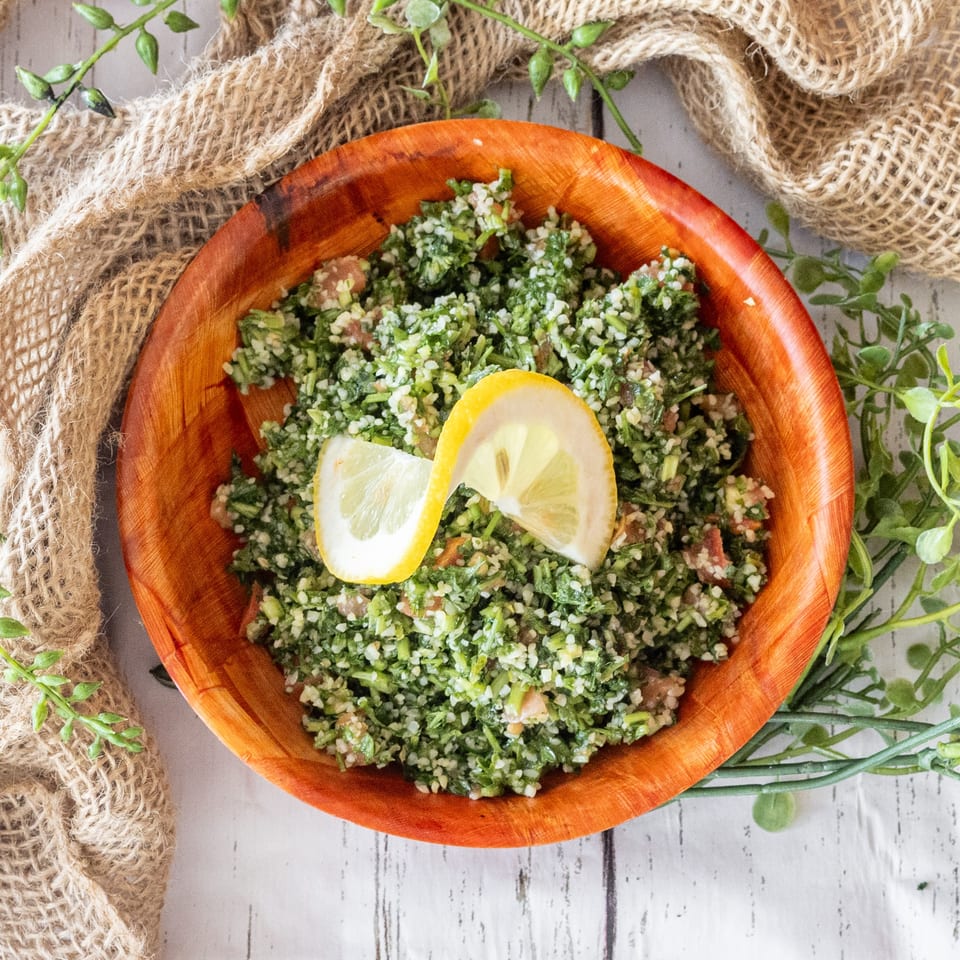A wooden bowl of vibrant green tabbouleh salad, garnished with two lemon slices, on a white wooden table.