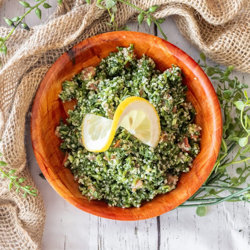 A wooden bowl of vibrant green tabbouleh salad, garnished with two lemon slices, on a white wooden table.