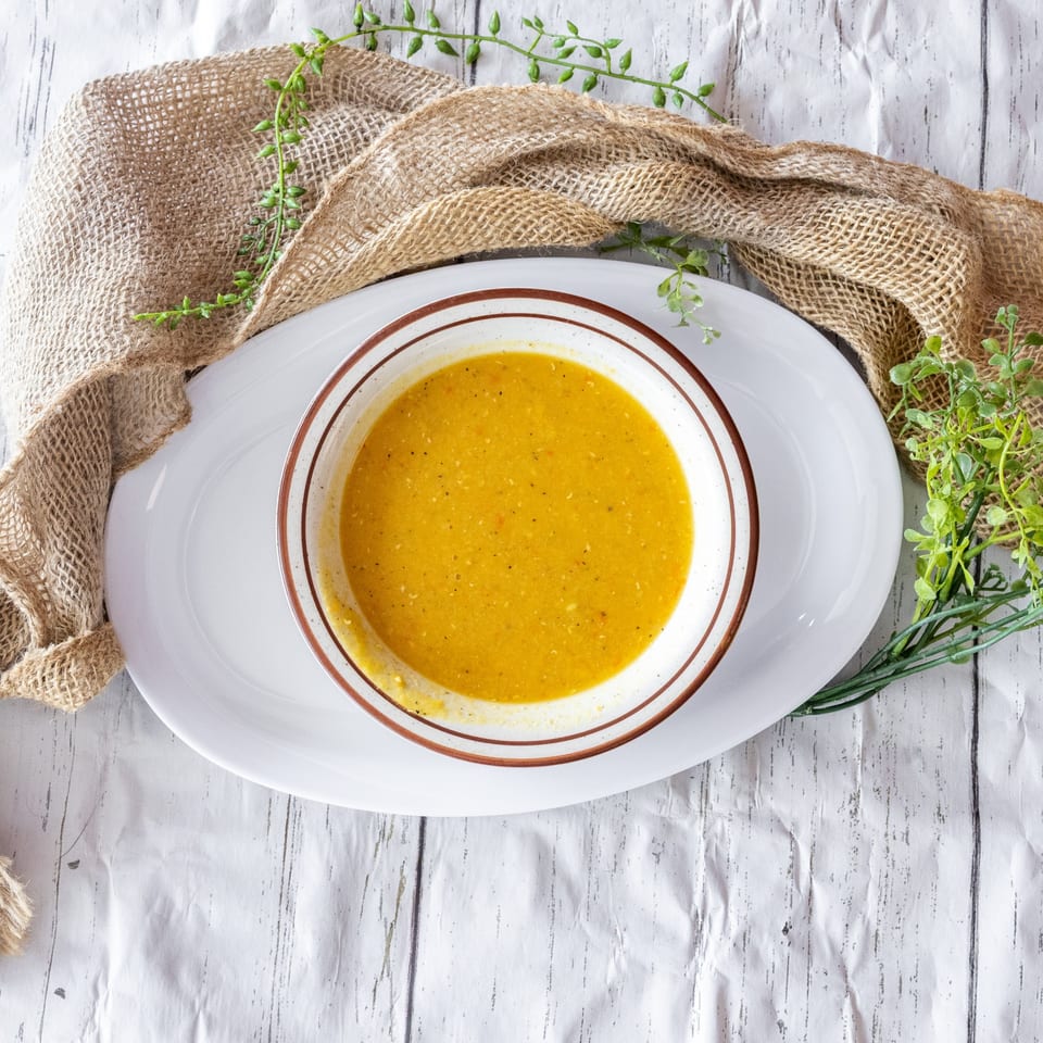 Overhead view of a bowl of yellow soup on a white platter with burlap and greenery.