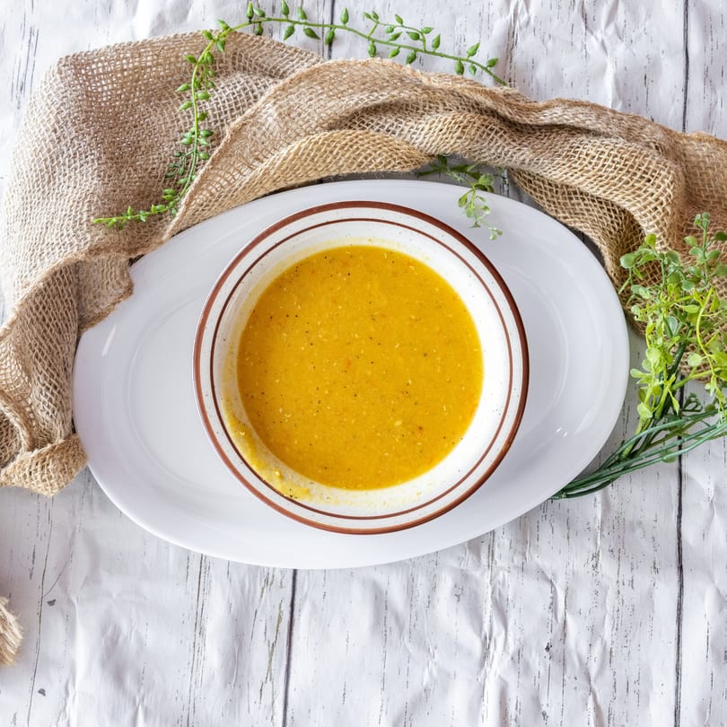 Overhead view of a bowl of yellow soup on a white platter with burlap and greenery.