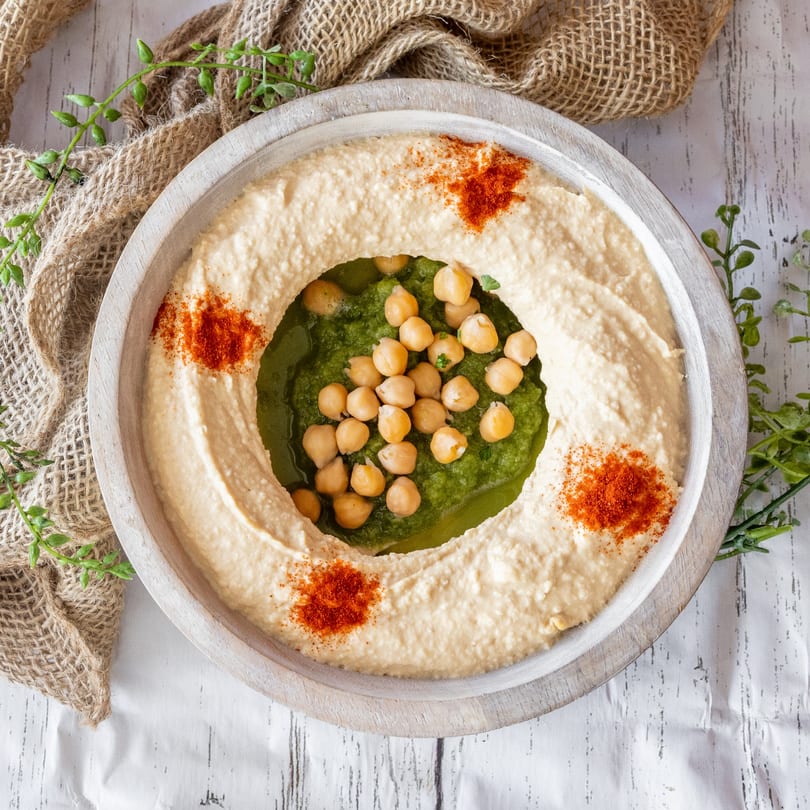 A wooden bowl of hummus with a green pesto center topped with chickpeas and paprika.