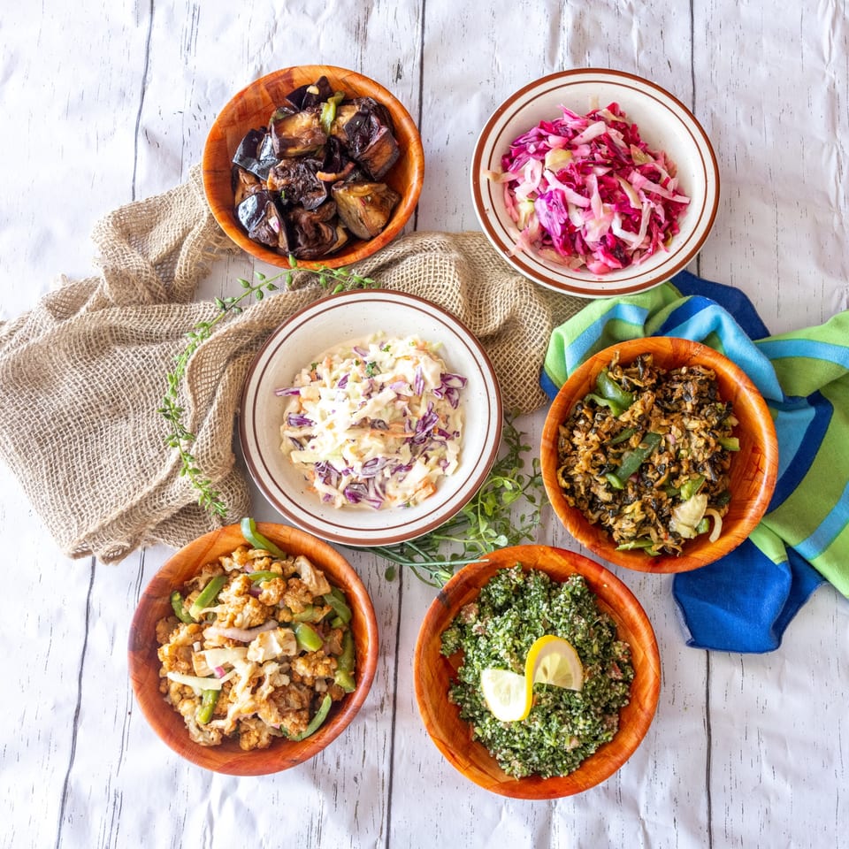 Overhead view of five wooden bowls filled with various salads and cooked vegetables on a white surface.