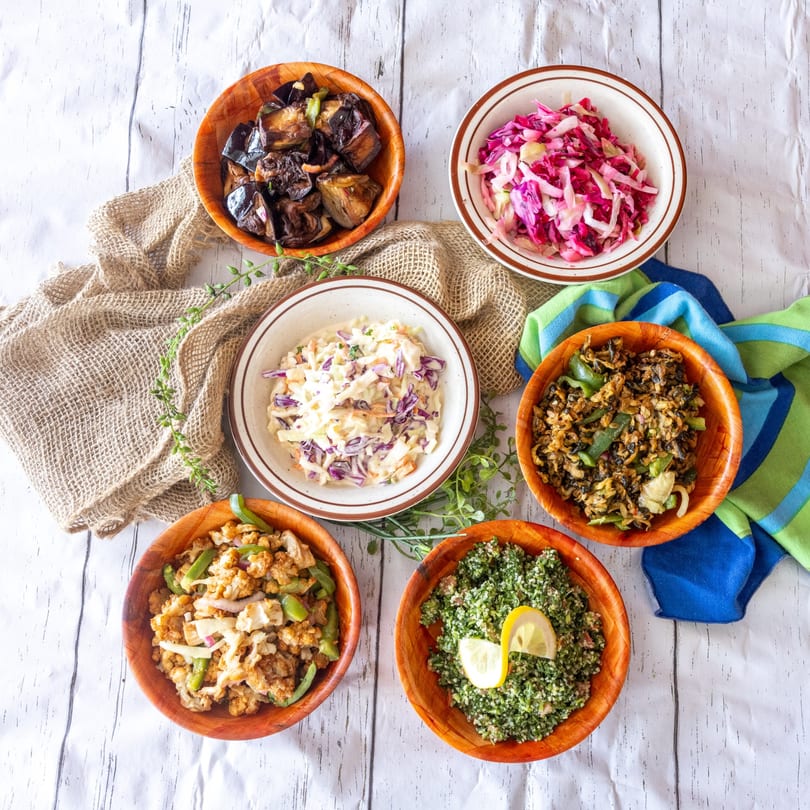 Overhead view of five wooden bowls filled with various salads and cooked vegetables on a white surface.