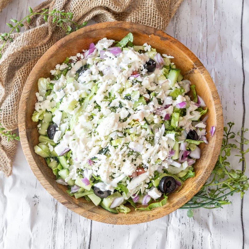 A wooden bowl filled with a fresh salad, topped with feta cheese, olives, and red onions.