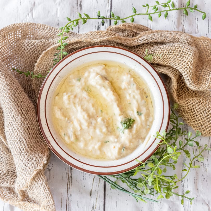 Creamy white dip with green herbs in a brown-rimmed bowl on a rustic white table with burlap.