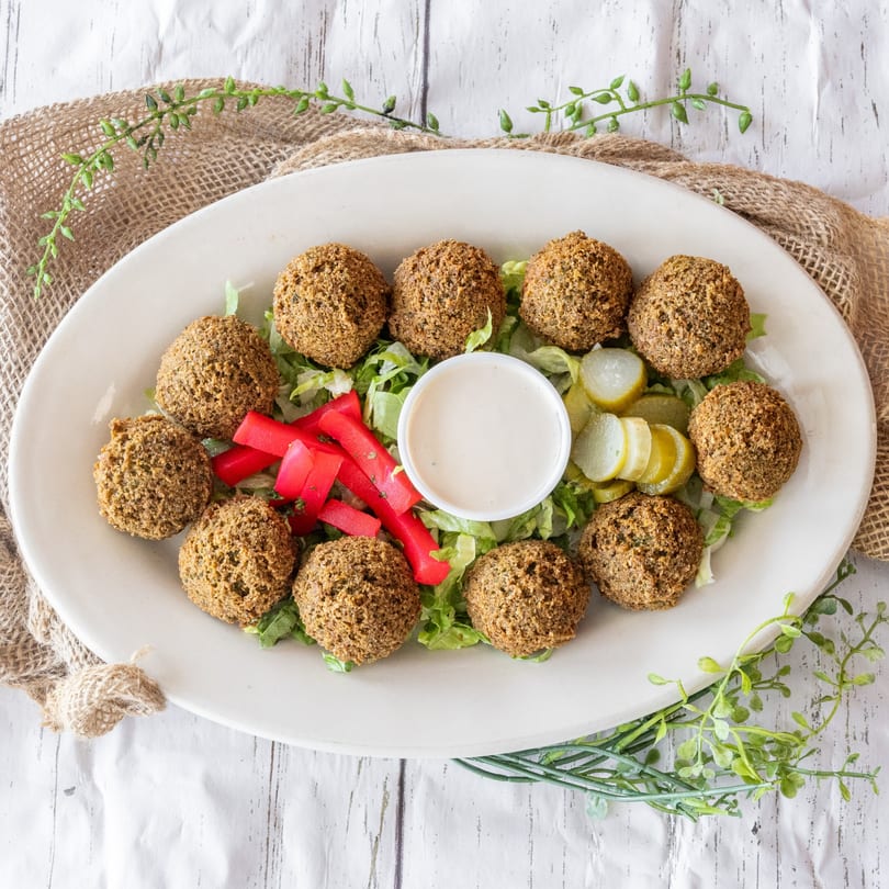 A platter of falafel balls on lettuce with pickled vegetables, red peppers, and a white sauce.