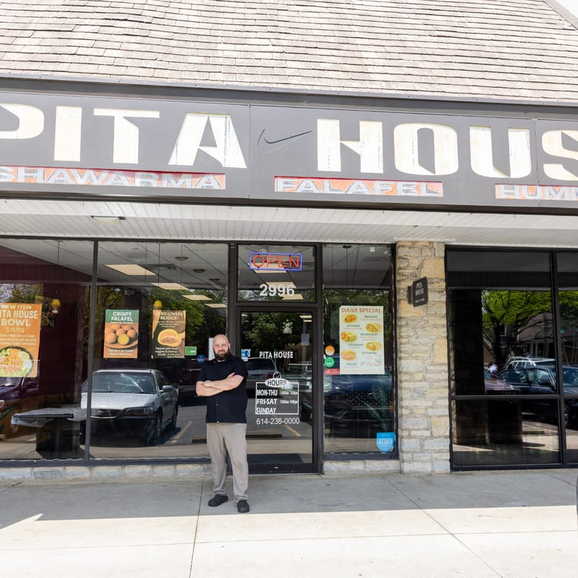 A man stands proudly in front of Pita House, a restaurant with a prominent sign and window advertisements.