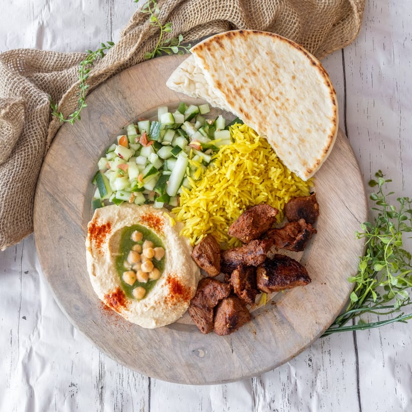 Mediterranean platter with hummus, pita, yellow rice, cucumber salad, and grilled meat on a wooden board.
